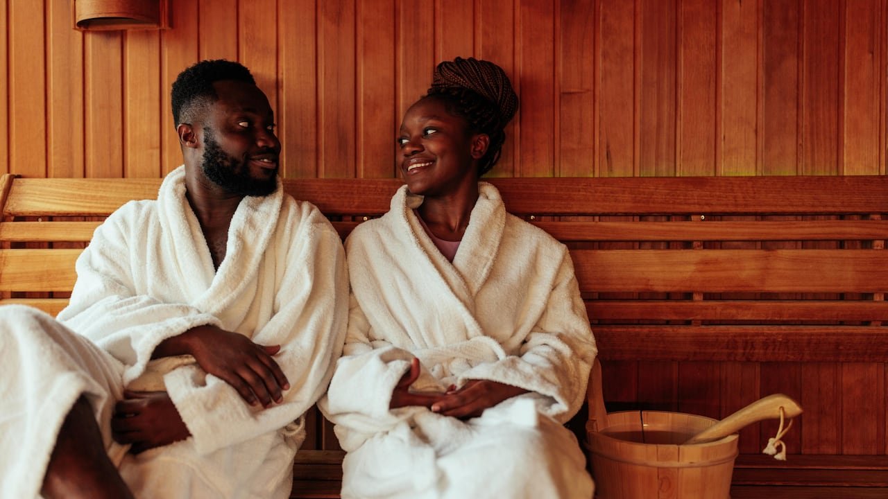 default-28 A couple in bath robes sitting in a wood-panelled sauna, smiling at each other.
