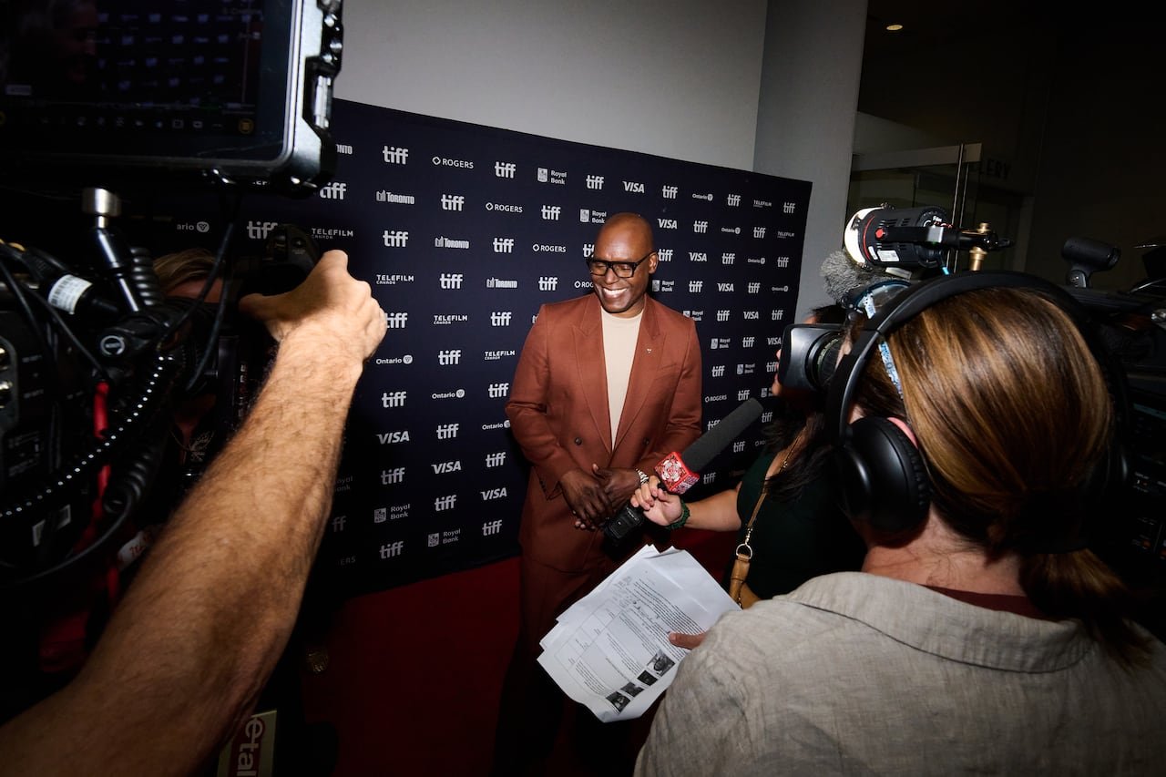 default-89 A man wearing a brown suit and glasses stands on the TIFF red carpet with cameras around him, with a black screen background behind him bearing the words "TIFF."
